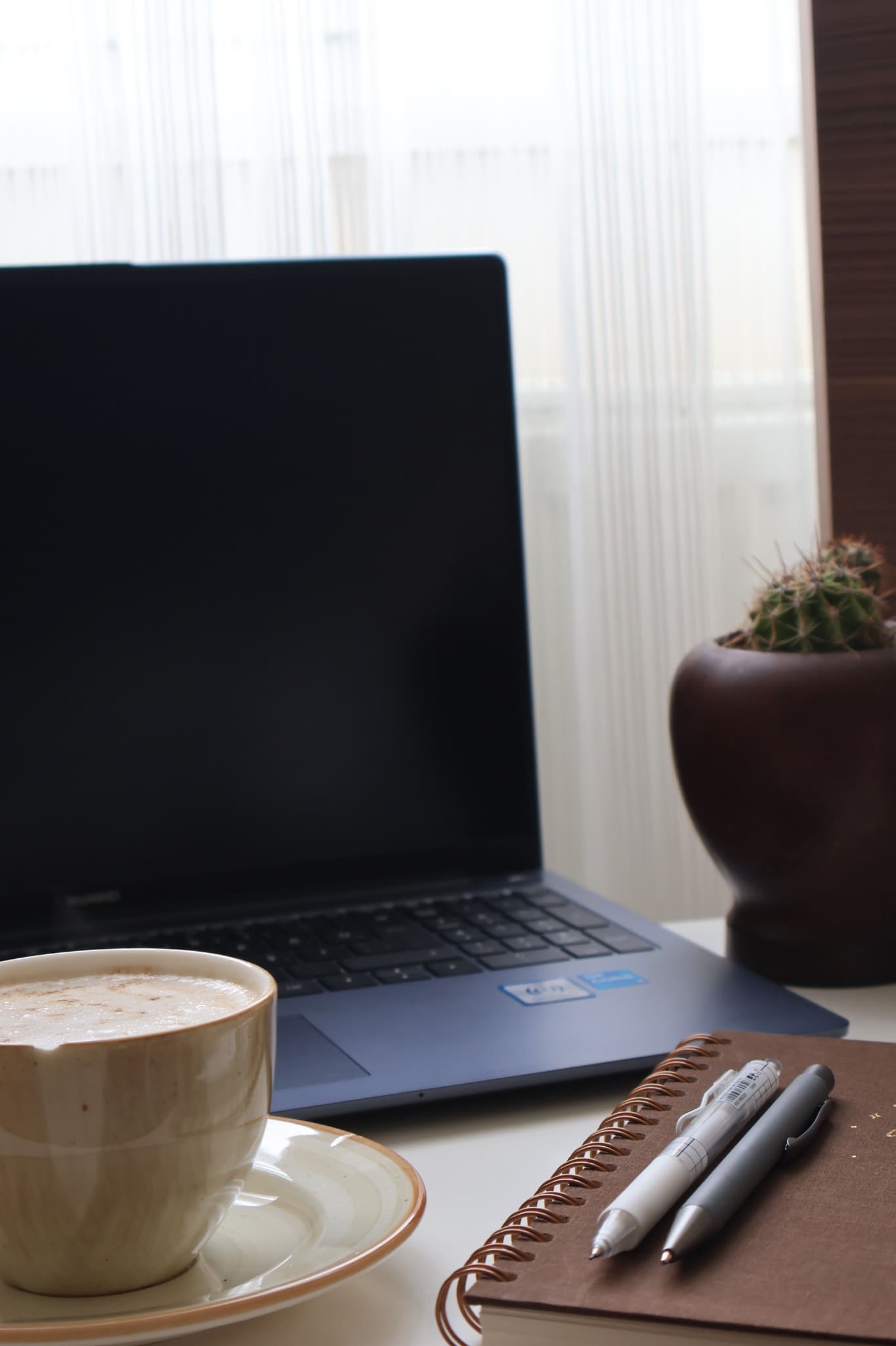 A quiet daylight workspace: an open laptop, a cappuccino, a spiral notebook with two pens and a small cactus on a wooden desk.